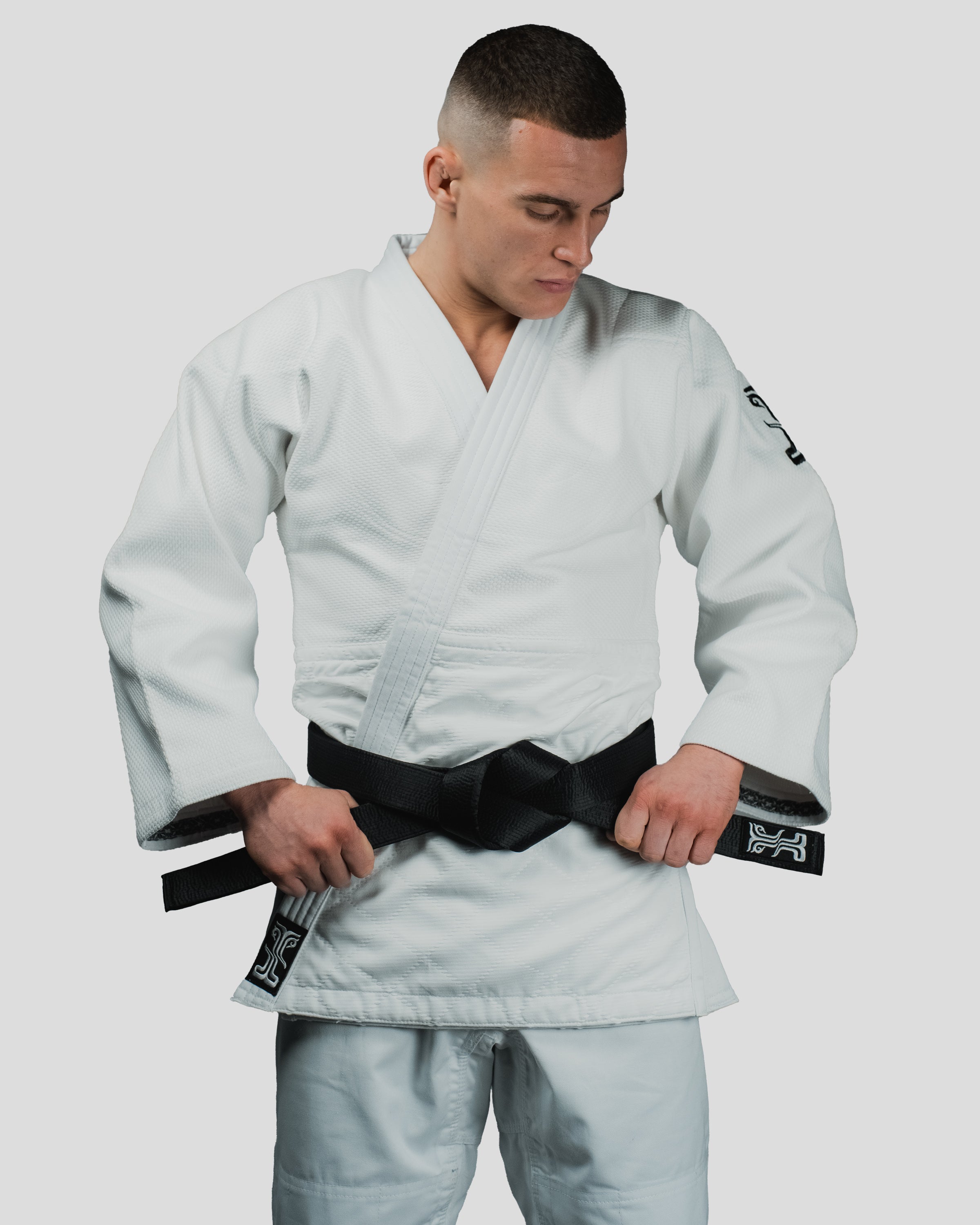 Martial artist tightening a black belt while wearing the Stoa Gi Men's Judo uniform, featuring the embroidered Kosmos logo