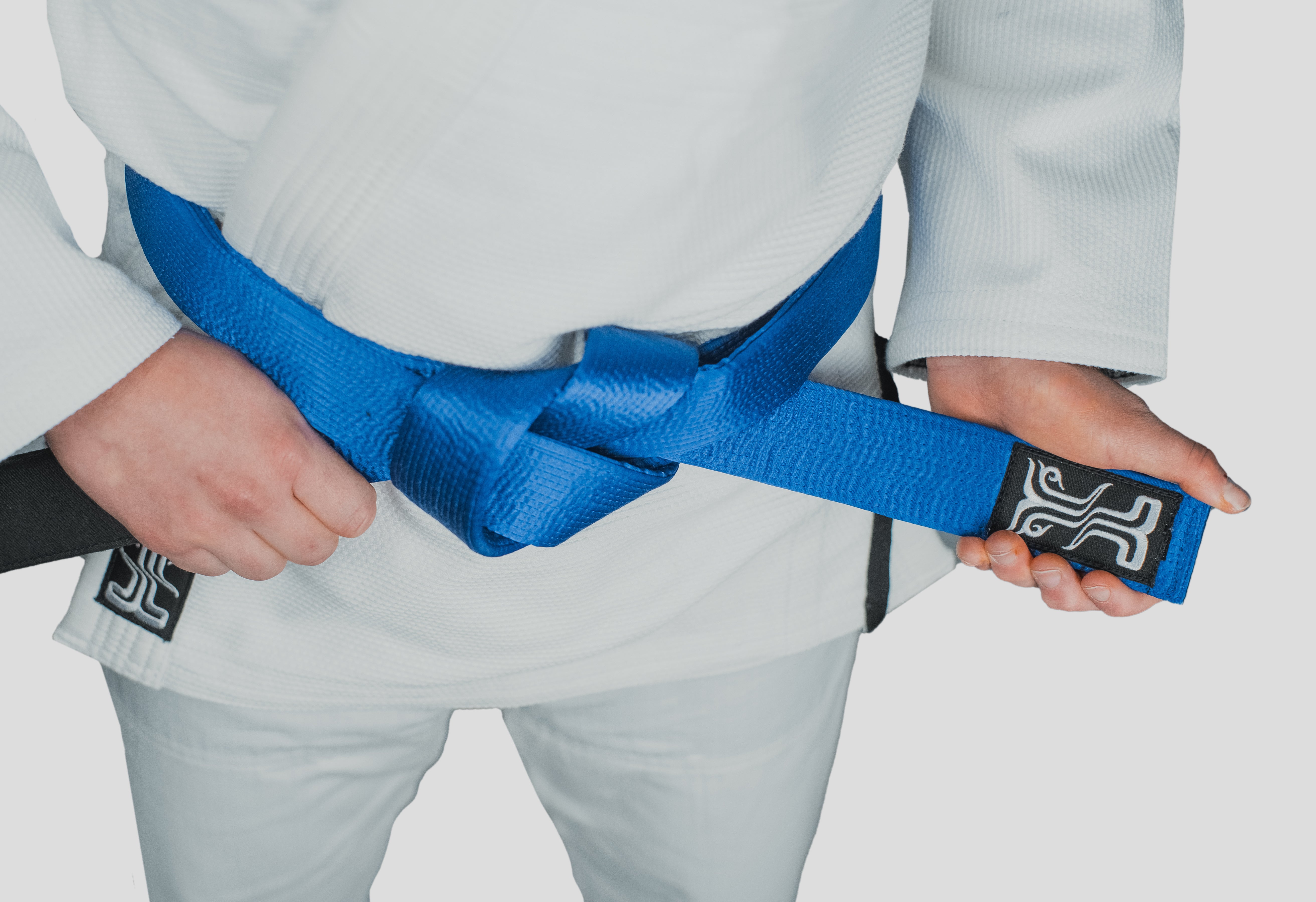 Close-up of the blue Tenacity Judo Belt, tied around a Kosmos gi, highlighting the embroidered logo and rank bar.